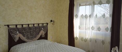 Un dormitorio con cama, mesita de noche, ventana con cortinas y lámpara fijada a la pared.