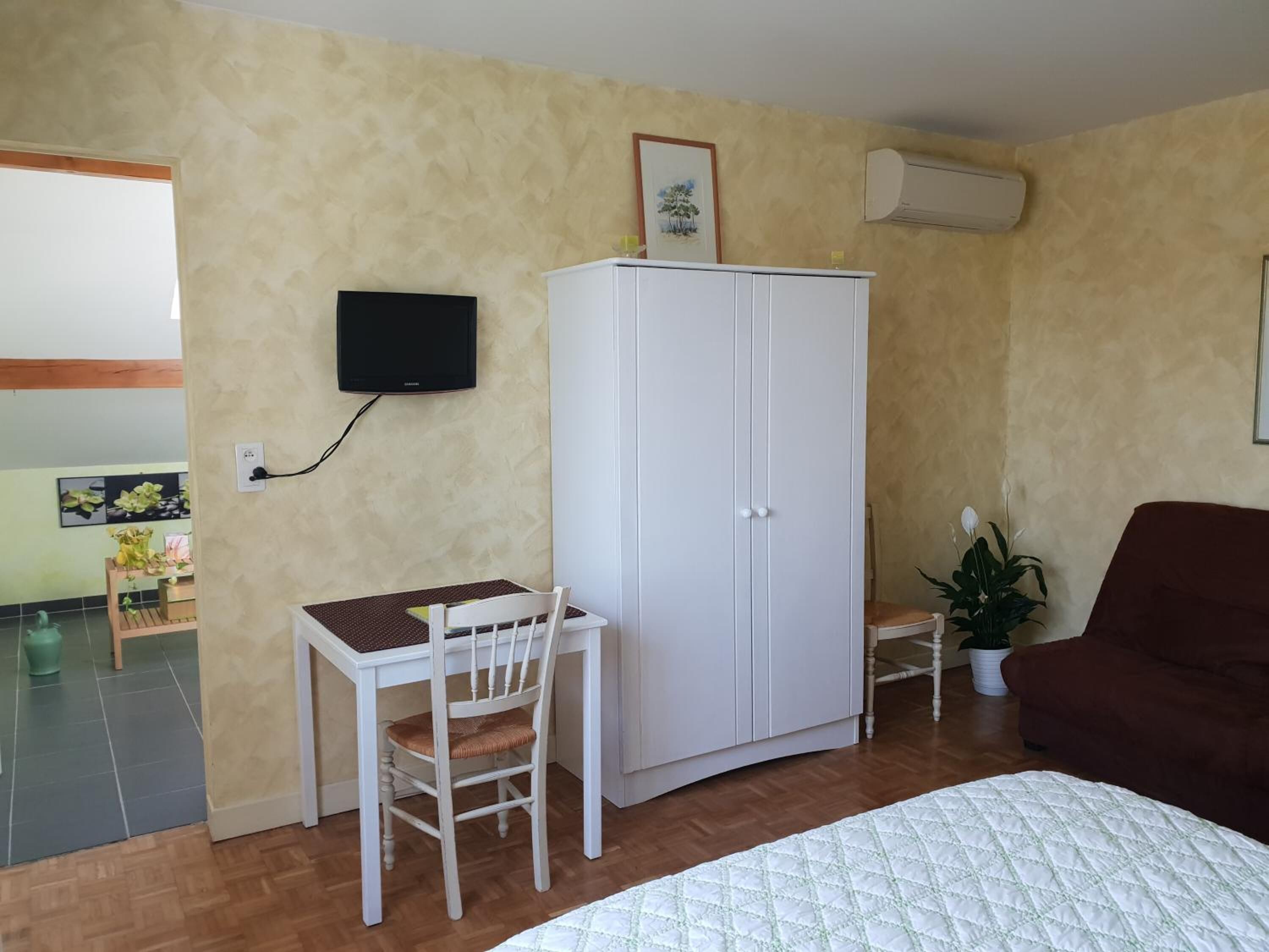 A bedroom with a bed, a desk with a chair, a wardrobe, a television, and a potted plant.