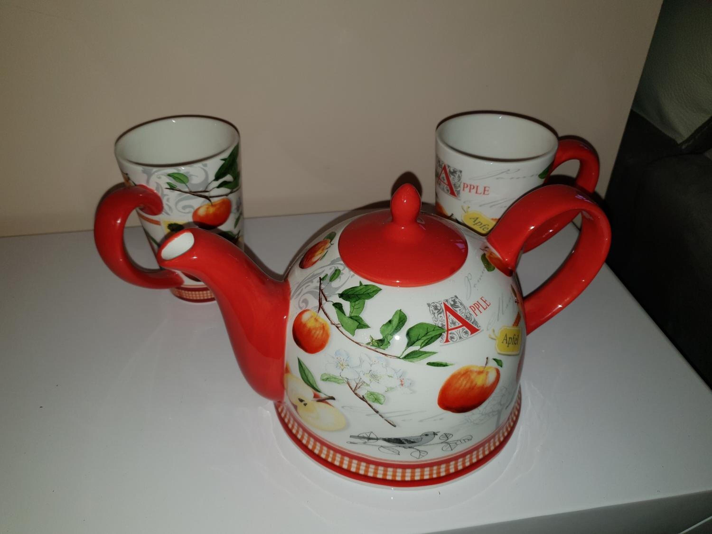 A white teapot with red handles and a matching cup set with apple designs.