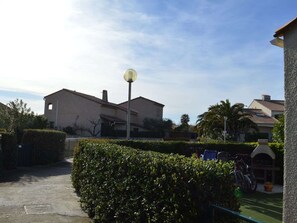 Ein Wohngebiet mit einem Weg, der zu einem Haus mit Ziegeldach und Schornstein führt. Im Vordergrund befindet sich eine Straßenlaterne und ein Heckenstreifen.