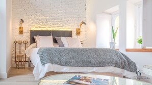Un dormitorio moderno con pared de ladrillo blanco, cama con ropa blanca, dos lámparas de noche y un taburete de madera.
