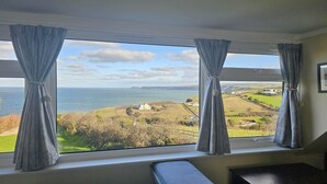 Ein Zimmer mit großem Fenster und Blick auf eine Küstenlandschaft, darunter ein Gewässer und ferne Hügel.