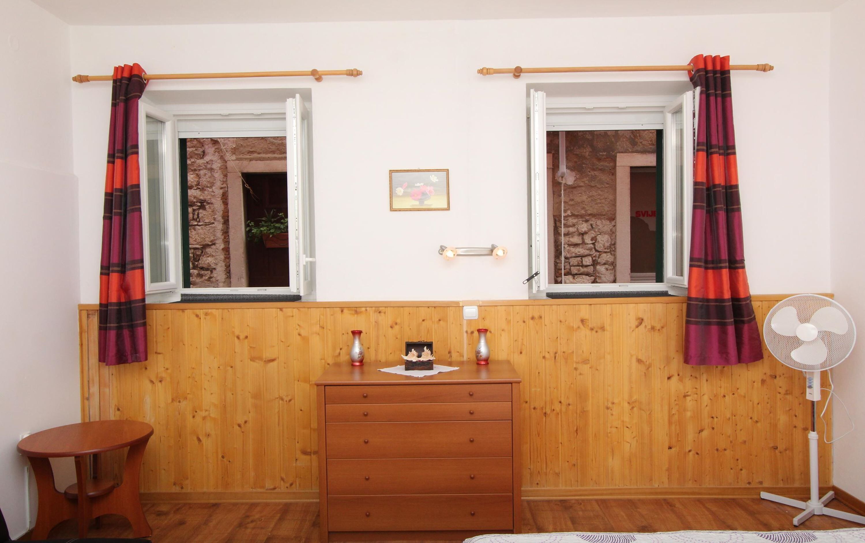 Una habitación con dos ventanas, paredes de madera, un aparador de madera, una mesita y un ventilador de pie.