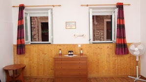A room with two windows, wooden walls, a wooden dresser, a small table, and a standing fan. - Studio Pupa (Rovinj)