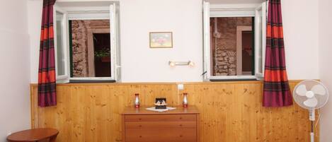 Una habitación con dos ventanas, paredes de madera, un aparador de madera, una mesita y un ventilador de pie.