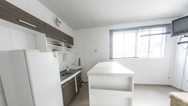 A modern kitchen with white cabinets, a built-in oven, and a window with blinds. - Bruno Henning - Apto 6 pessoas (Itapema)