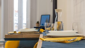 Ein Esstisch ist gedeckt mit Tellern, Gläsern und Besteck, im Hintergrund stehen ein Laptop und eine kleine Pflanze.