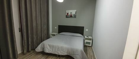Un dormitorio con cama, mesita de noche, luz en la pared y una puerta con cortinas.