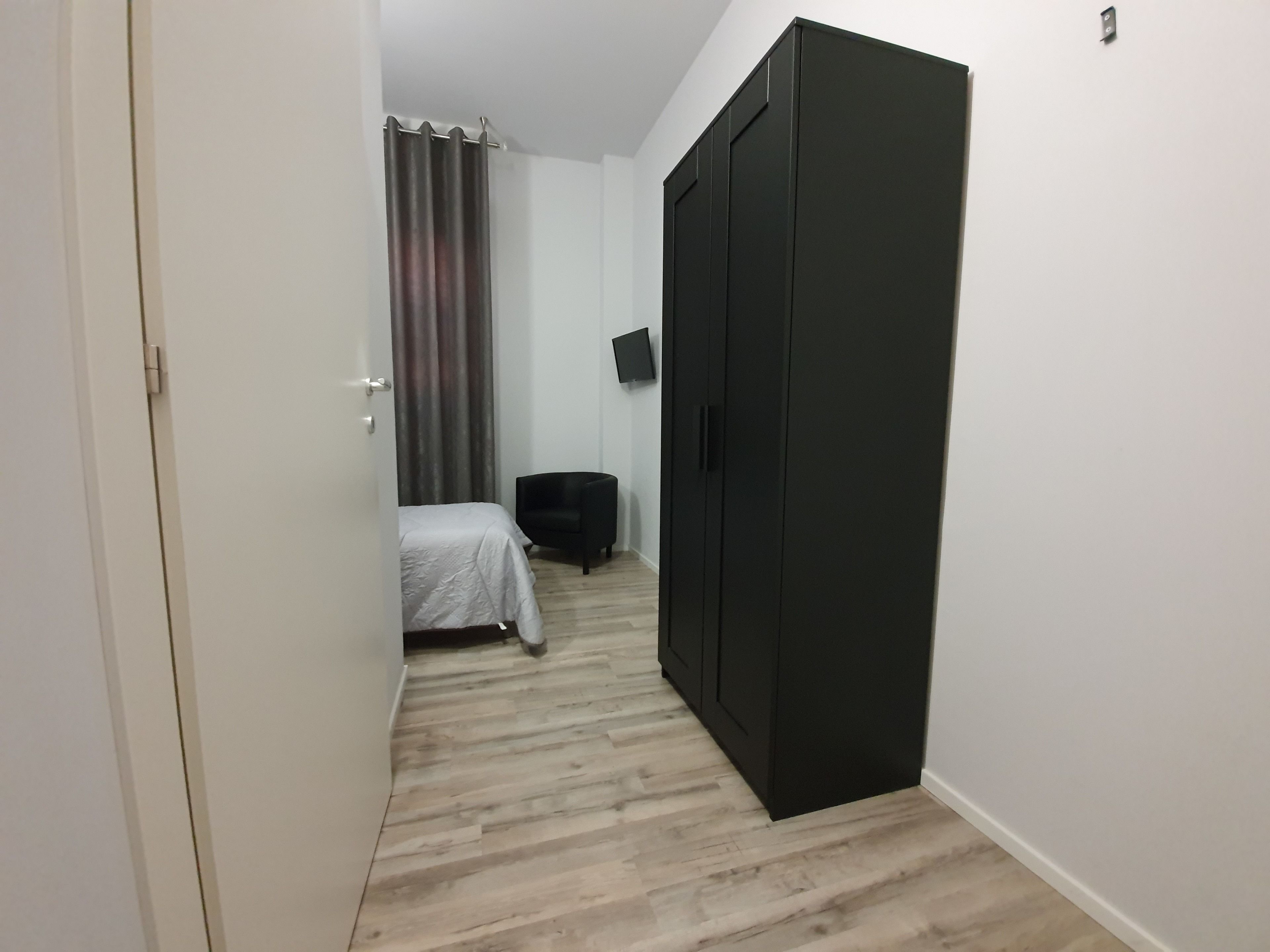 A narrow hallway with a bed, a black wardrobe, and a chair.