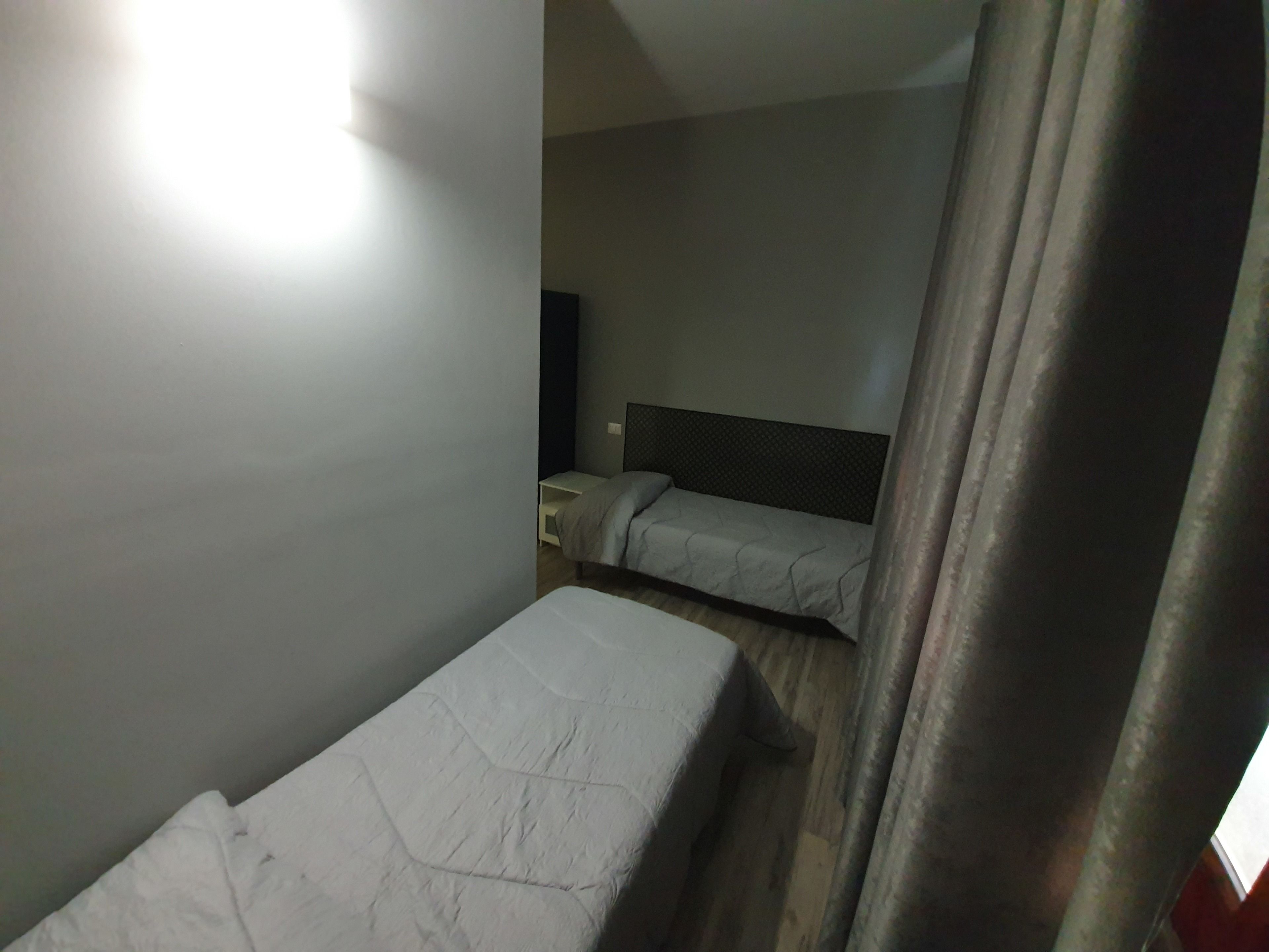 Una piccola stanza poco illuminata con due letti singoli, un comodino e una TV a muro.