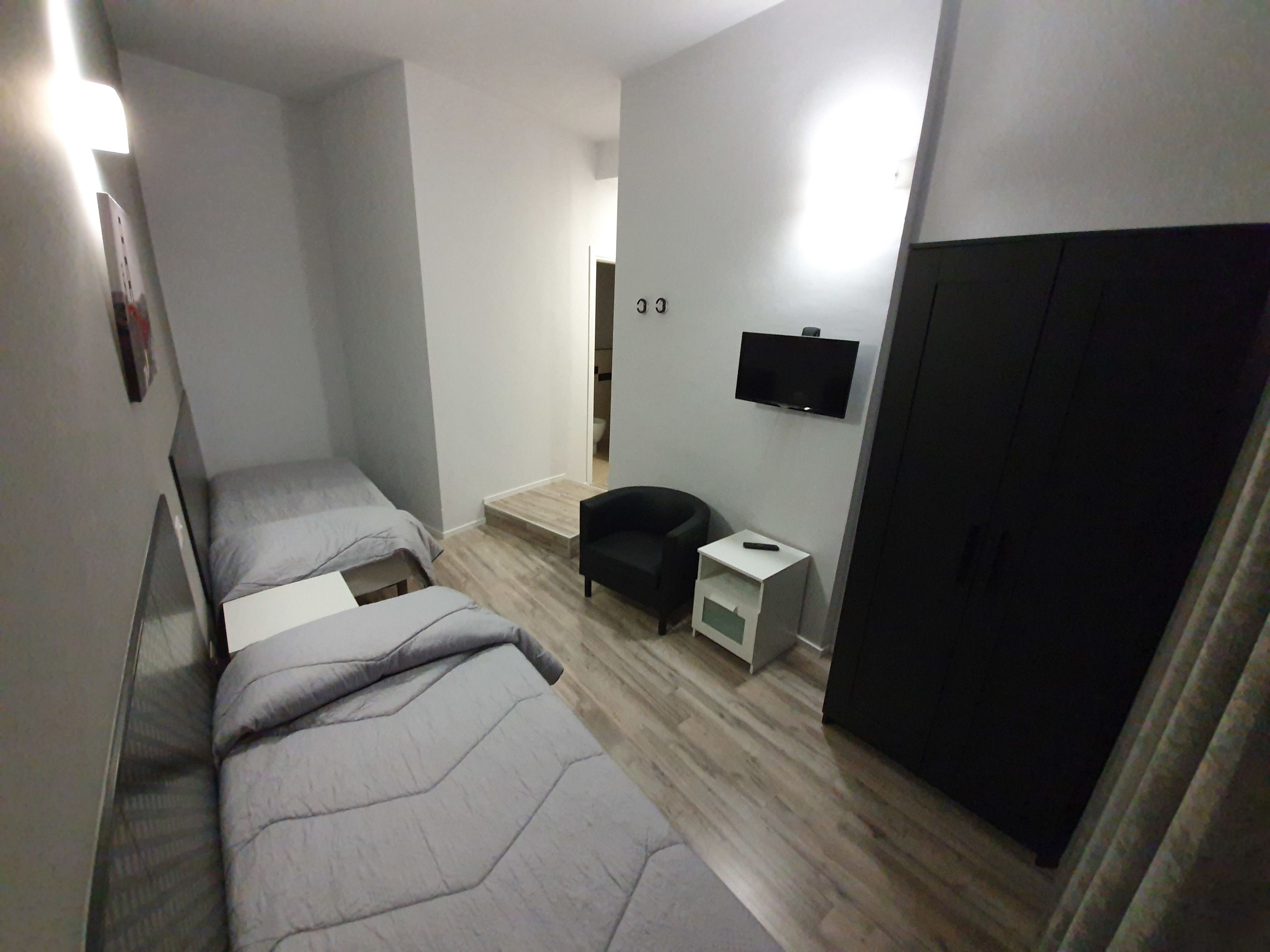 A compact room with two single beds, a small seating area, a TV mounted on the wall, and a wardrobe.