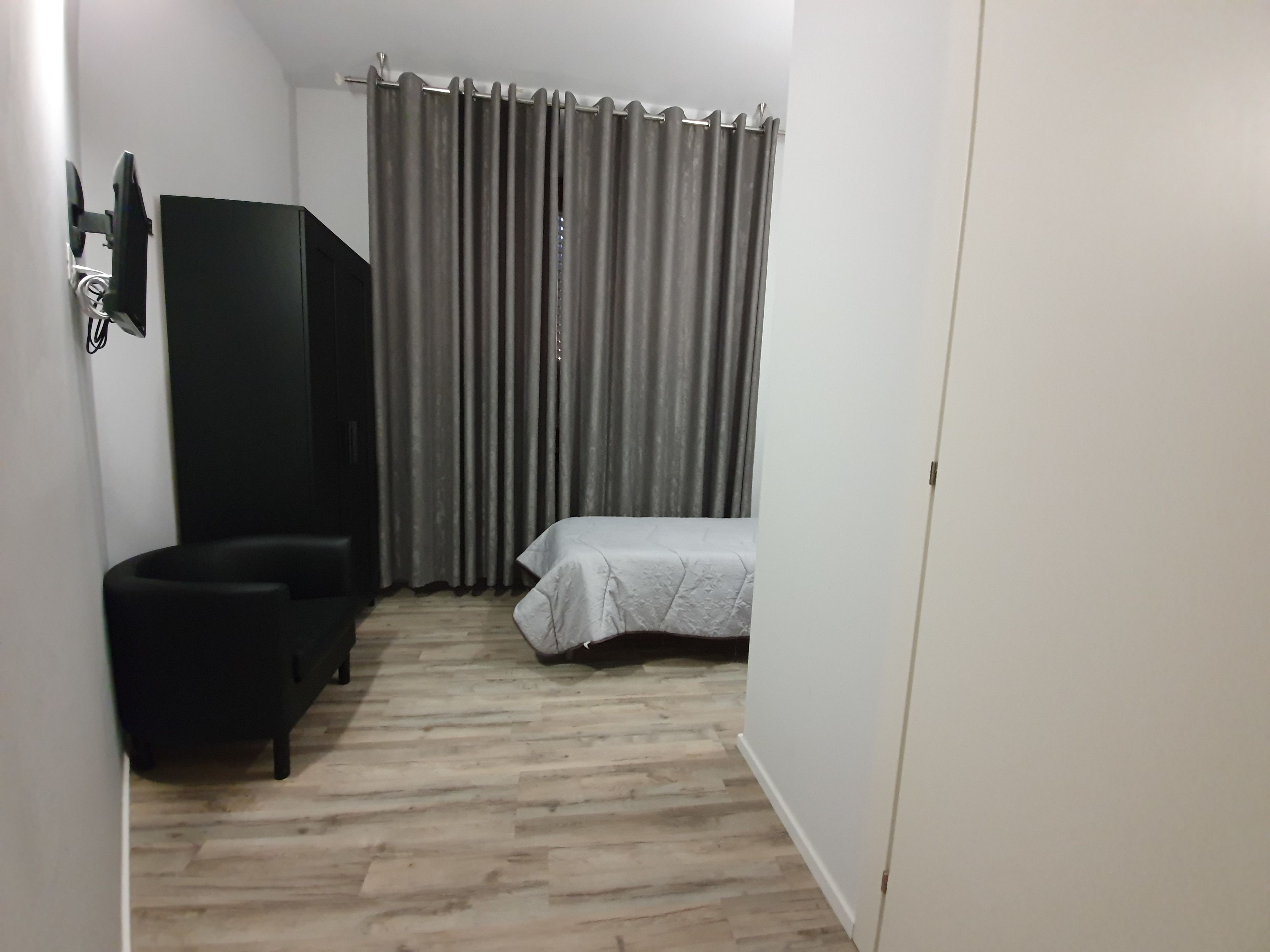 Um quarto pequeno, com pouca luz, com uma cama, uma cadeira e uma televisão na parede.
