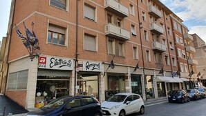 A multi-story building with a brick facade, featuring several ground-floor shops including an electrician and a tailor. - Affittacamere Umberto I (Foligno)