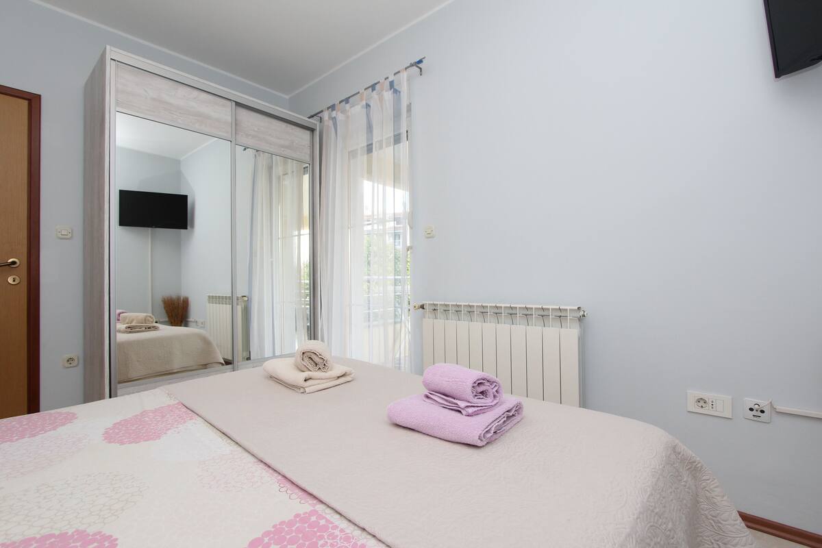 Un dormitorio con cama, espejo, televisor y ventana con cortinas.