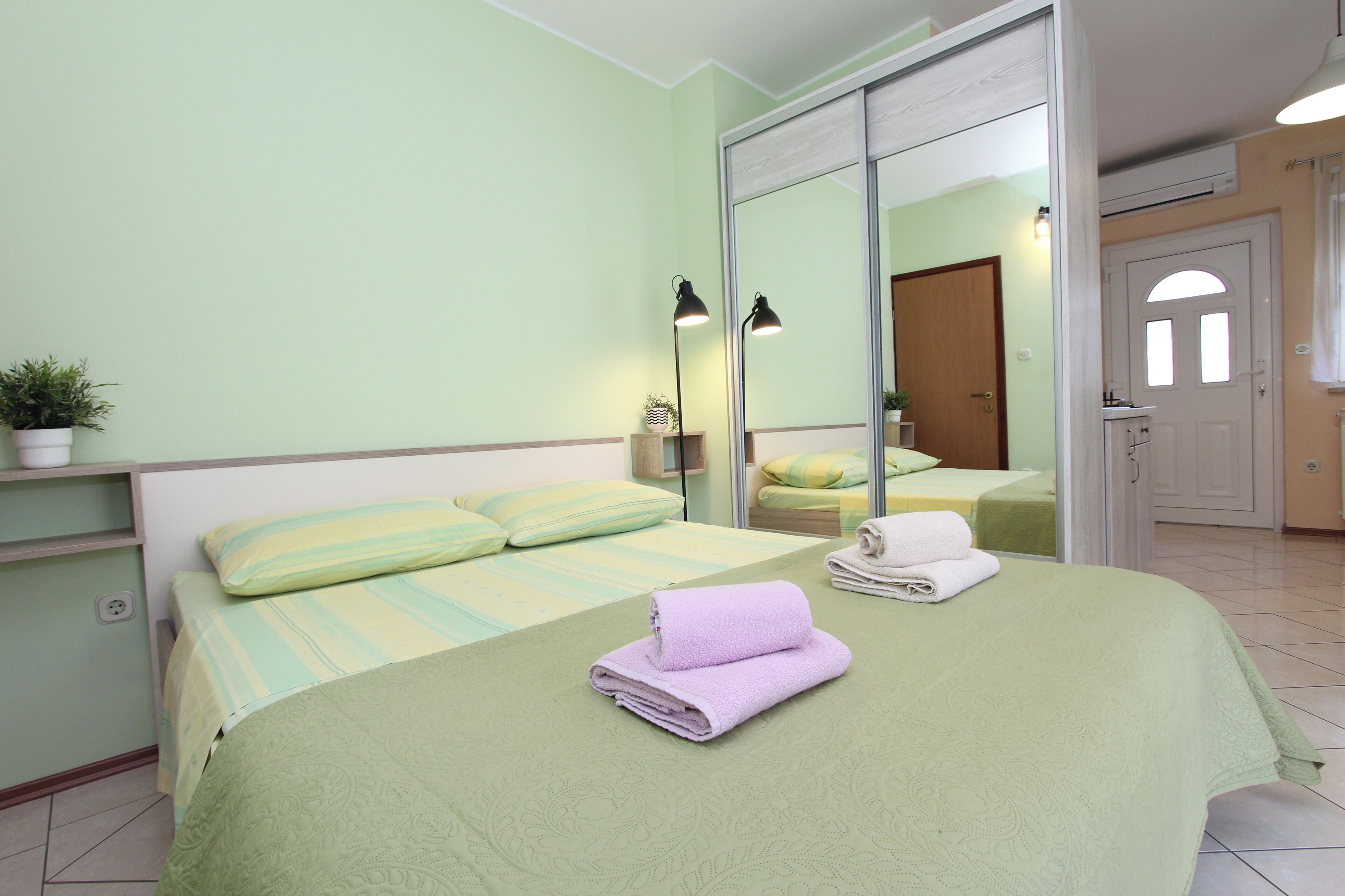 A bedroom with a bed, a mirror, a shelf with a plant, and a door leading to another room.