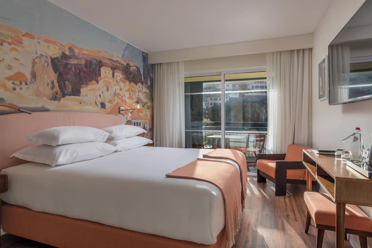 A hotel room with a large bed, a desk, a chair, and a view of the city through the window.