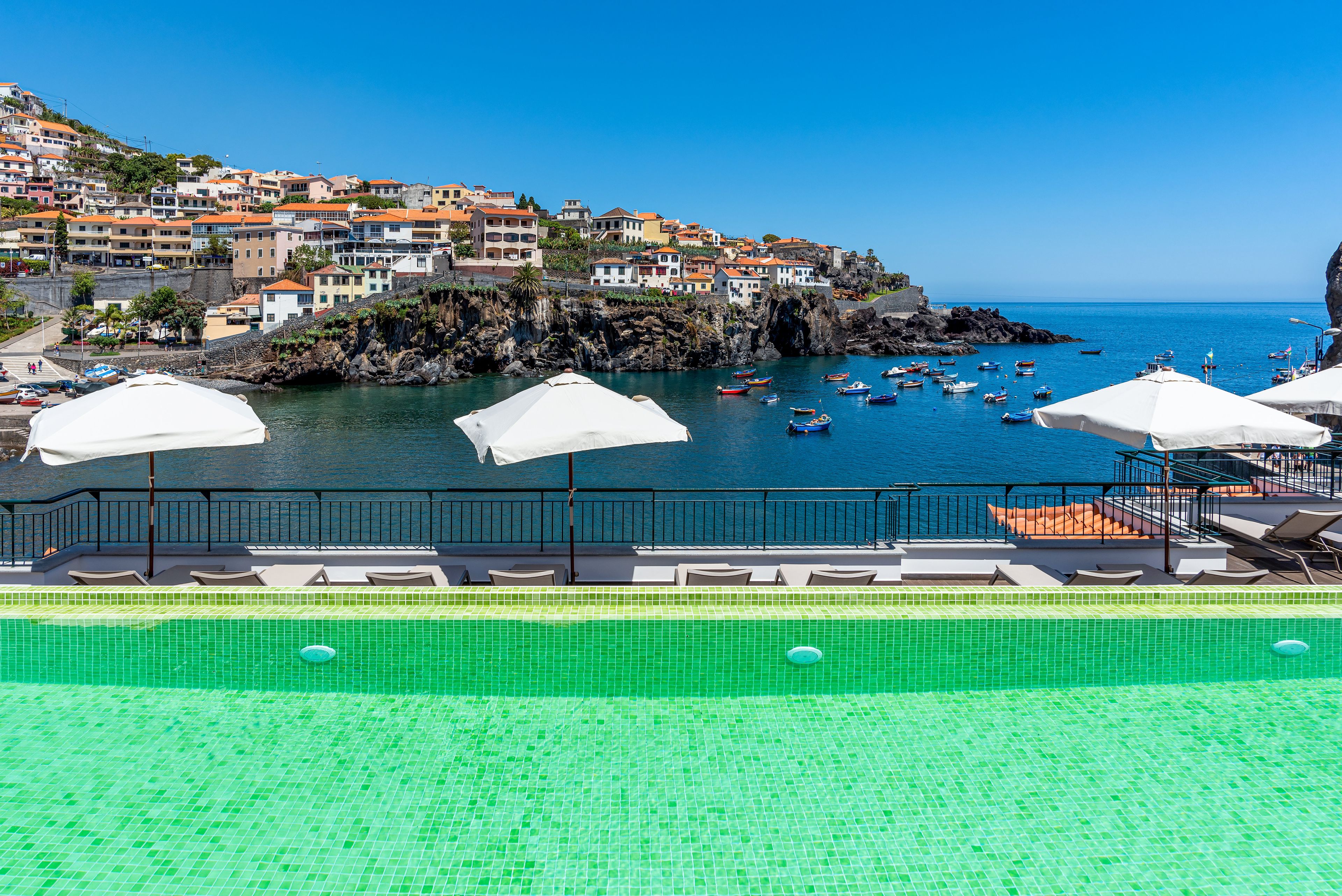 Un espace aménagé au bord de la piscine offrant une vue sur une ville côtière, avec notamment un littoral rocheux et des bateaux sur l’eau.