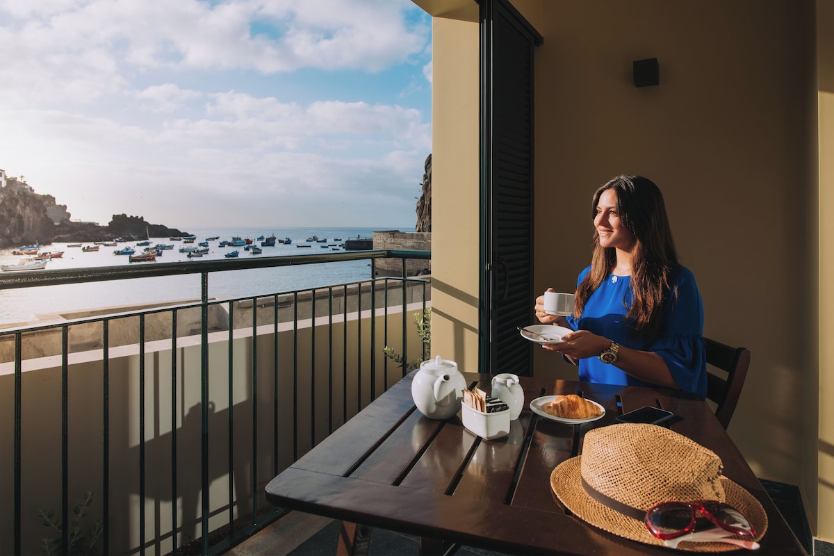 A balcony with a table set for breakfast, overlooking a coastal area with boats and cliffs.