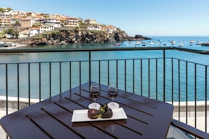 A table with a plate of food and two glasses of red wine, overlooking a coastal town with boats in the water.