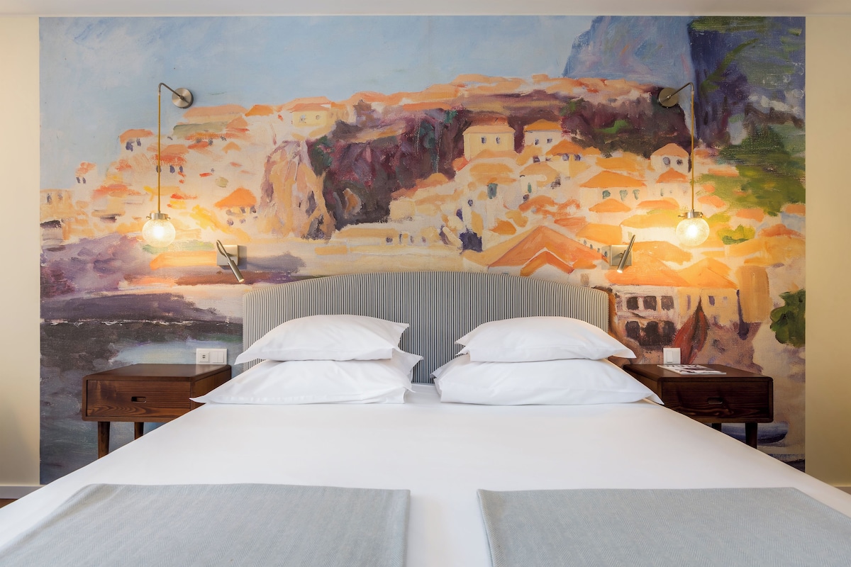 A hotel room with a bed, two bedside tables, and a large painting on the wall.