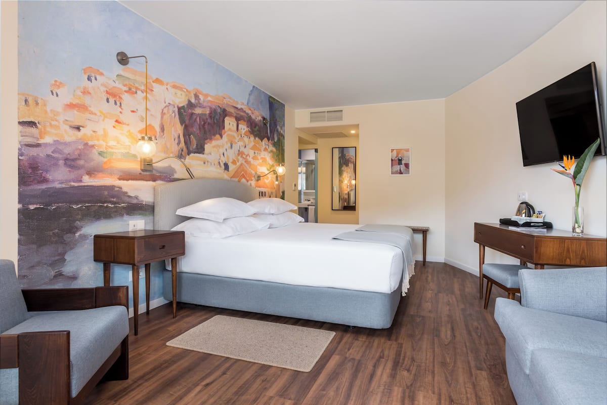 A modern hotel room with a large bed, a desk, a chair, a TV, and a painting on the wall.