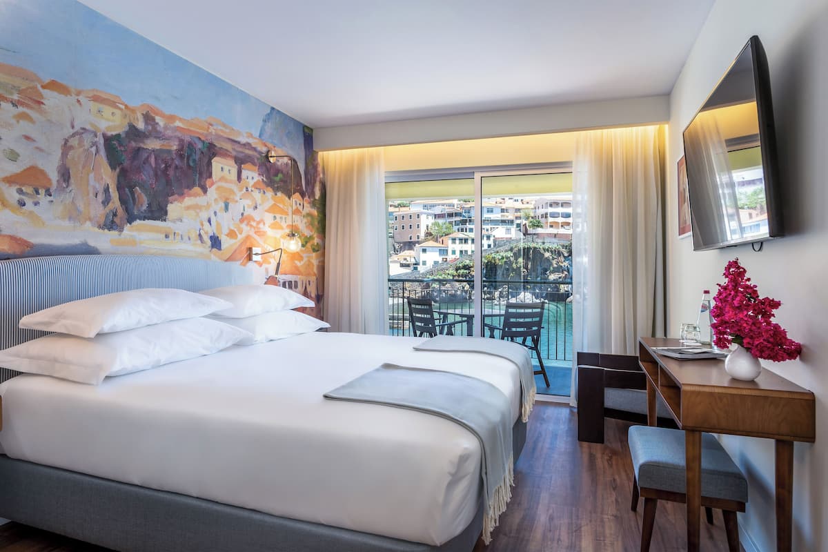 A hotel room with a large bed, a desk with a chair, a balcony with a view of buildings, and a painting on the wall.