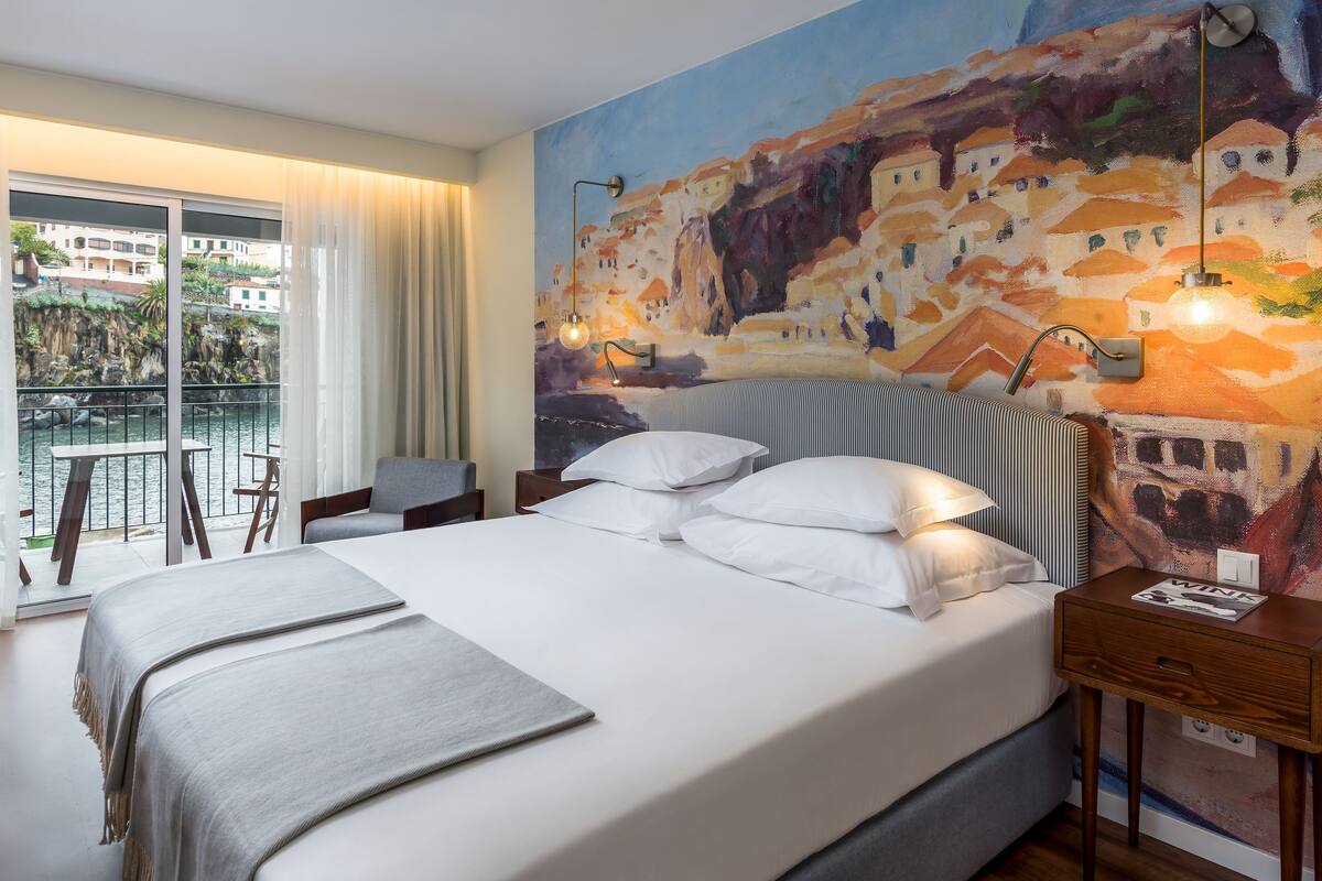 A hotel room with a bed, a nightstand, a chair, a table, a balcony with a view of buildings and water.