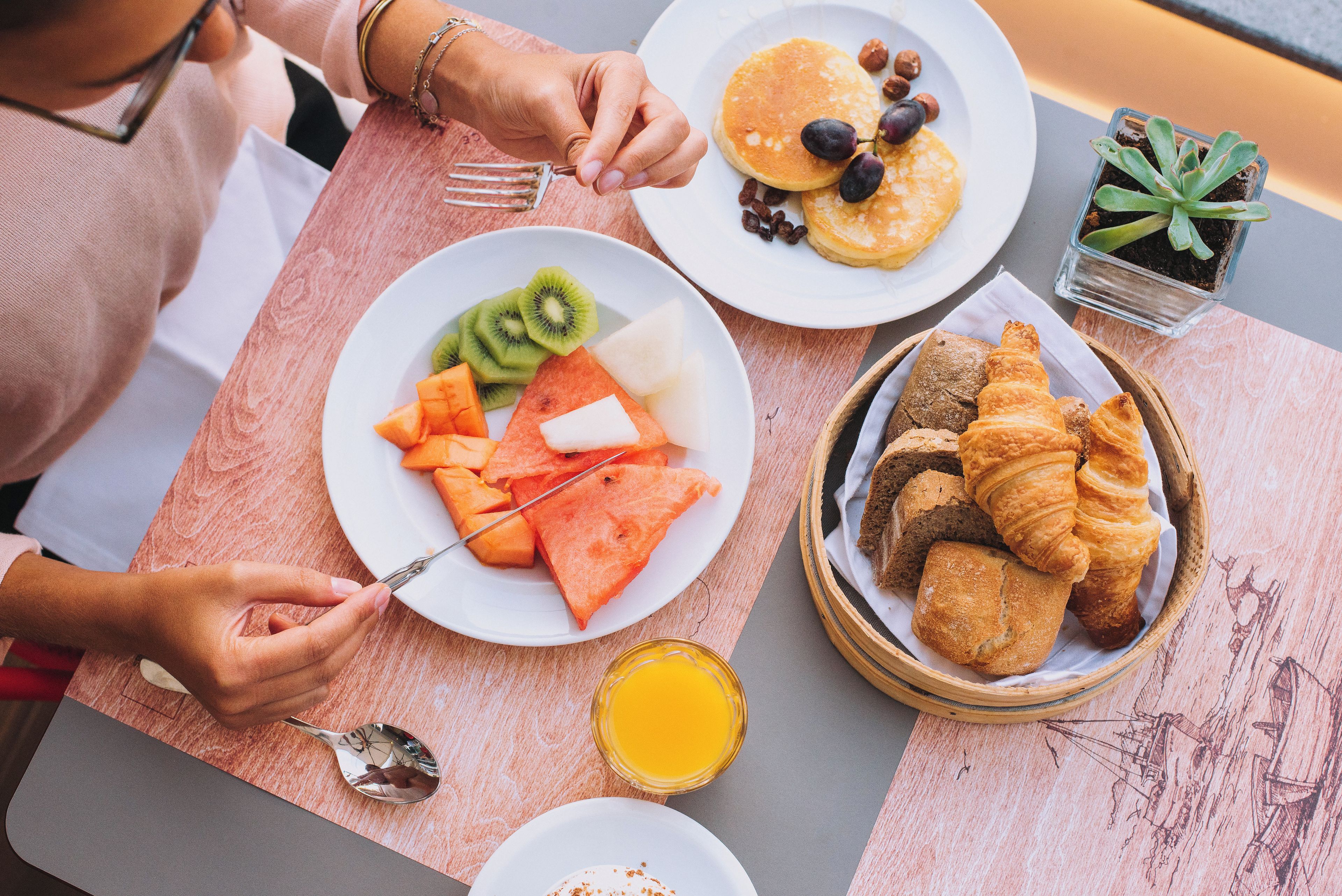A breakfast setting with plates of food, a basket of croissants, and a small potted plant.