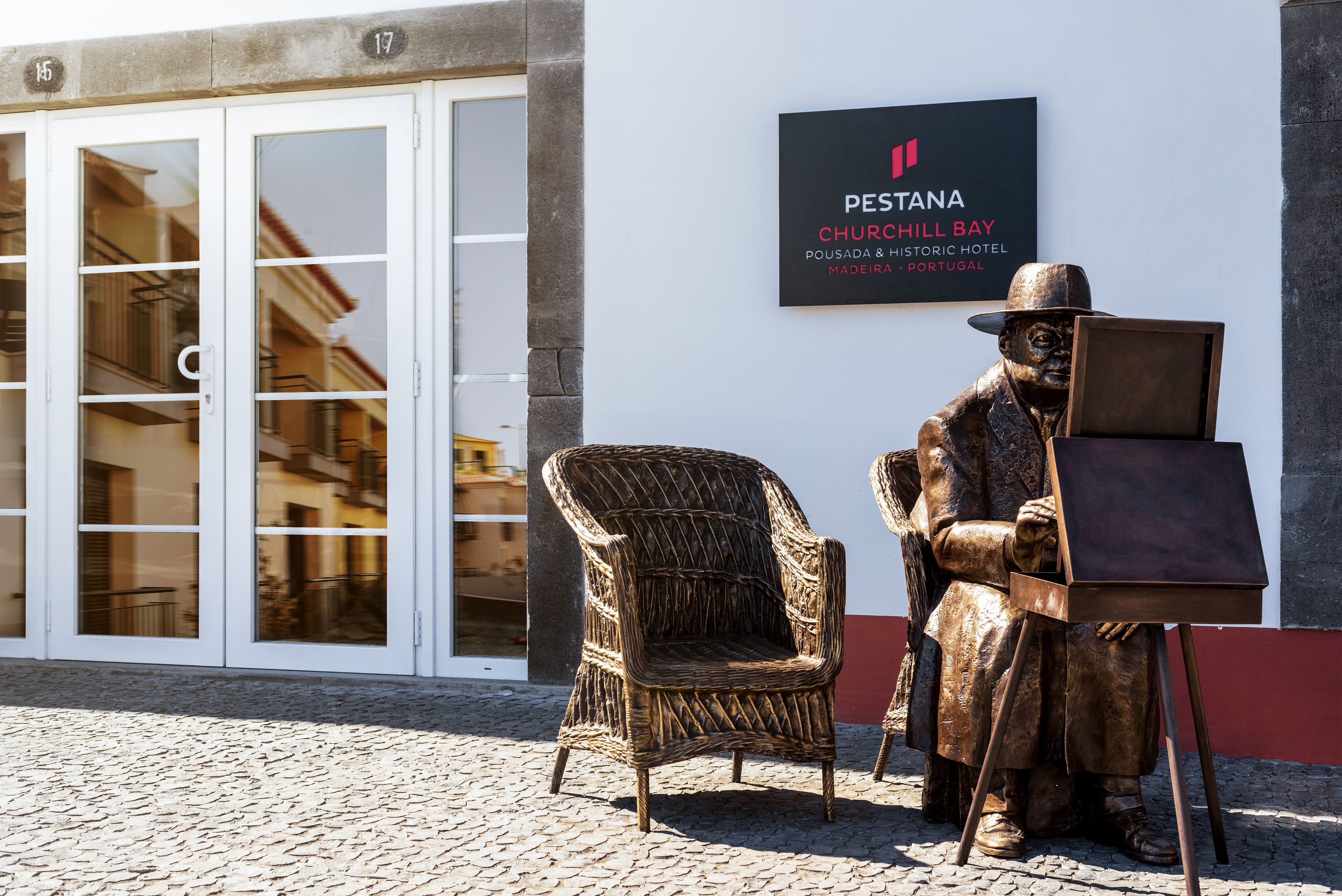 Een standbeeld van een zittende man met een schilderij op een voetstuk, een rieten stoel, en een bordje voor PESTANA CHURCHILL BAY POUSADA & HISTORIC HOTEL in Madeira, Portugal.