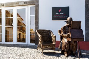 Una statua di un uomo seduto con un dipinto su un cavalletto, una sedia in vimini e un cartello per PESTANA CHURCHILL BAY POUSADA & HISTORIC HOTEL in Madeira, Portogallo.