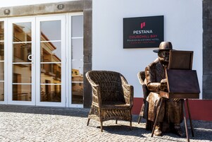 A statue of a seated man with a painting on a stand, a wicker chair, and a sign for PESTANA CHURCHILL BAY POUSADA & HISTORIC HOTEL in Madeira, Portugal.