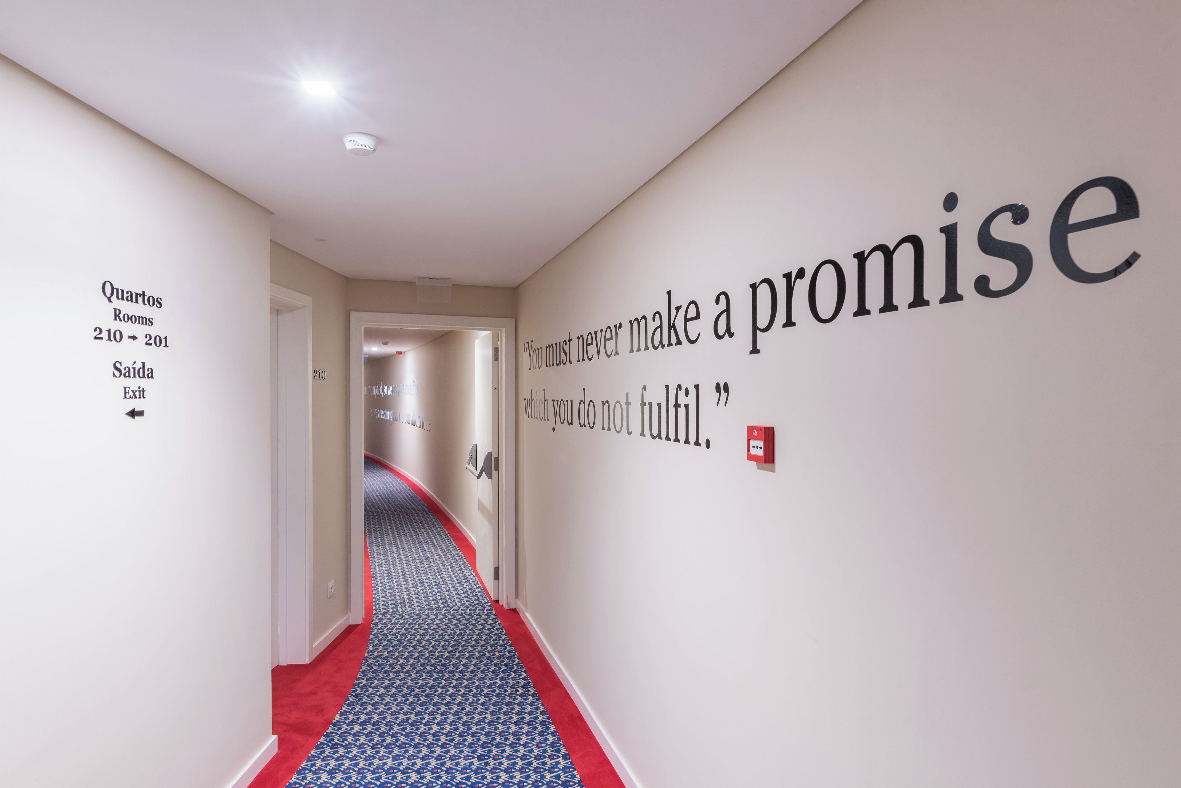 Ένας διάδρομος με κόκκινο χαλί, ένας τοίχος με ένα απόσπασμα και μια πινακίδα που υποδεικνύει τους αριθμούς των δωματίων 210 έως 201.