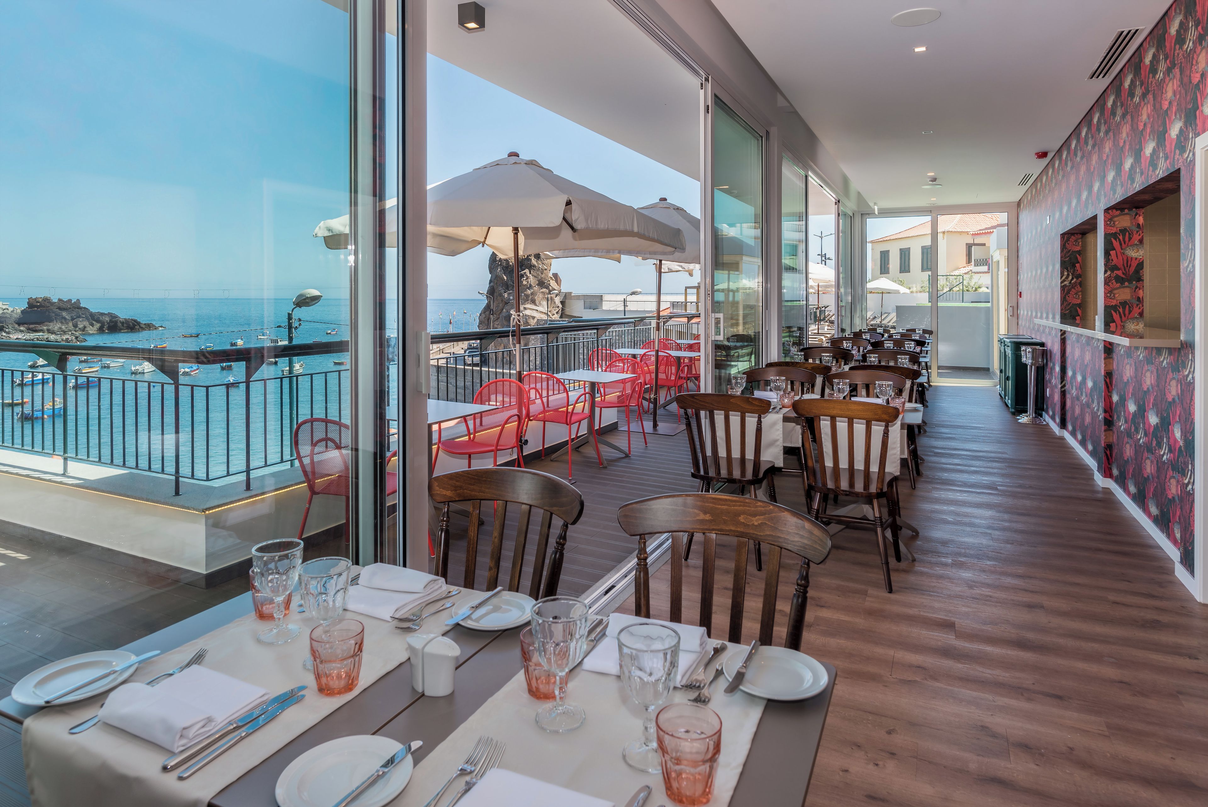A restaurant with tables set for dining, overlooking a rocky coastline with boats.
