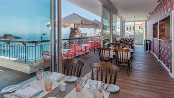 A restaurant with tables set for dining, overlooking a rocky coastline with boats.