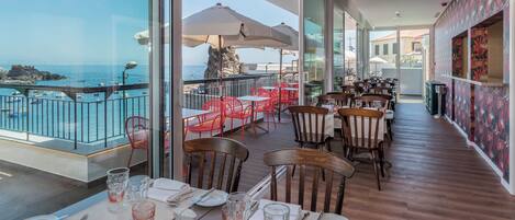 Un restaurante con mesas dispuestas para comer, con vista a una costa rocosa y barcos.