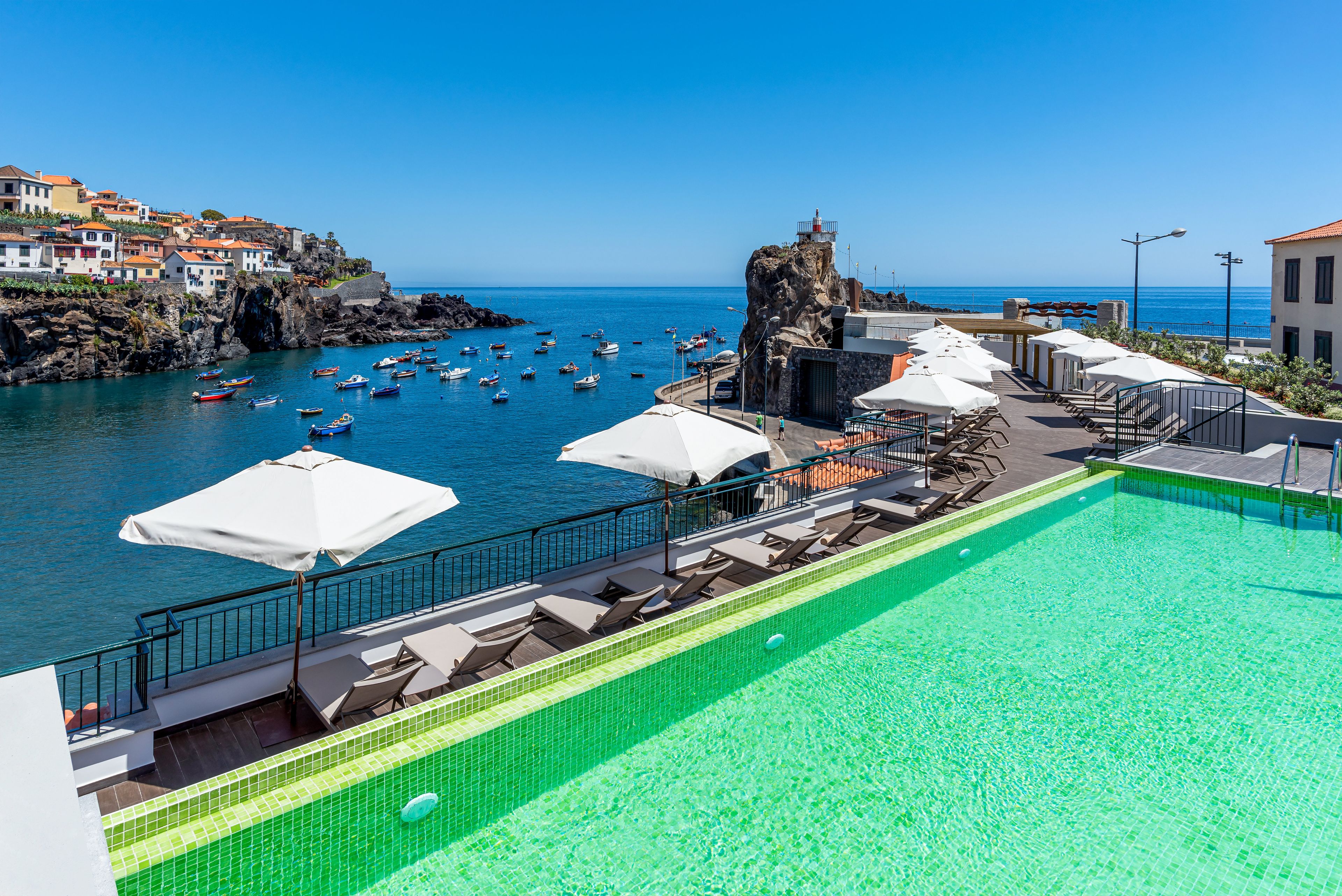 A rooftop pool with clear blue water, overlooking a rocky coastline with boats and buildings.
