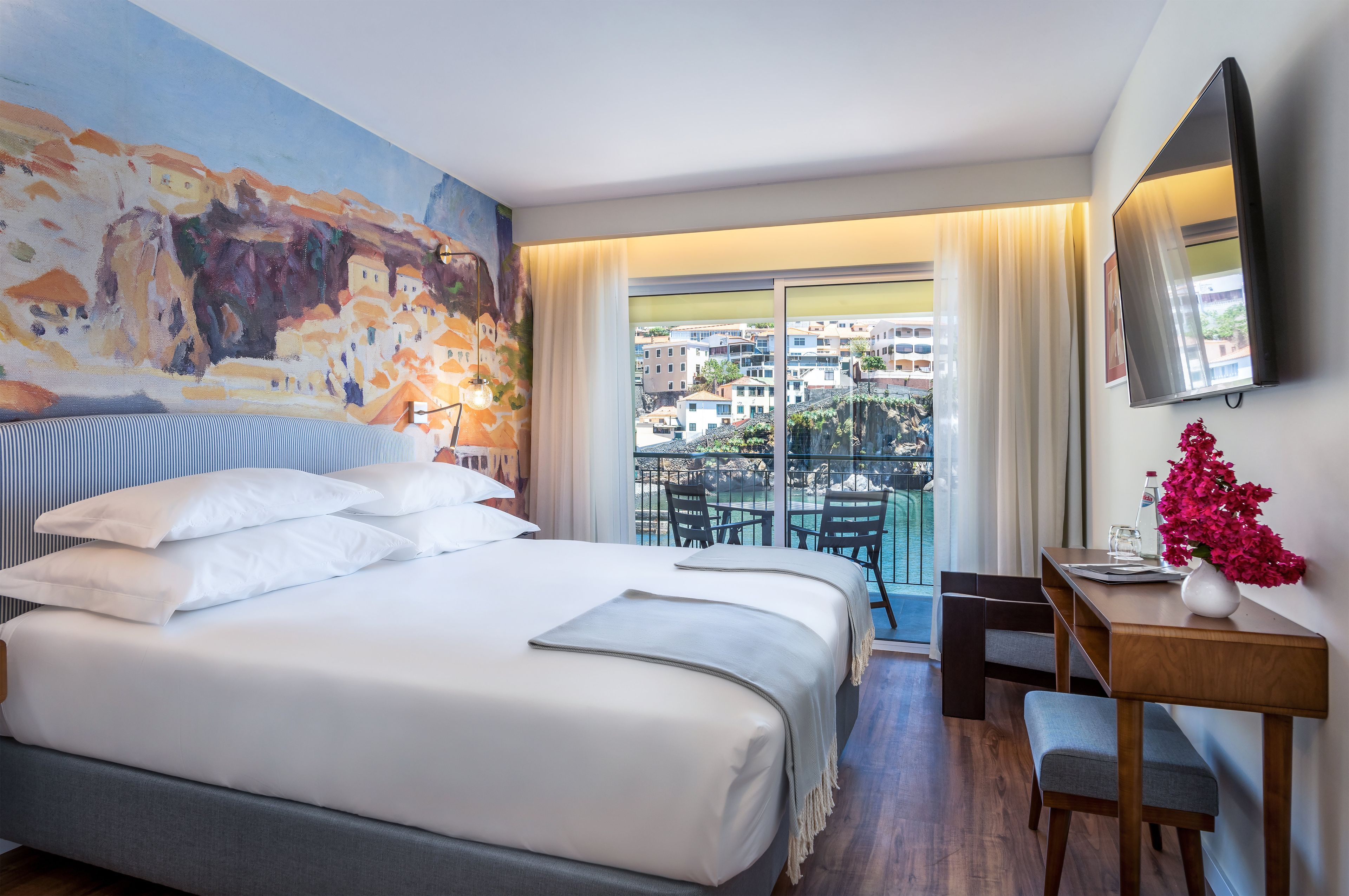 Ein Hotelzimmer mit Bett, Schreibtisch, Stuhl, einem Bild an der Wand und einem Balkon mit Blick auf Gebäude.