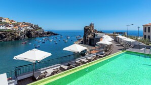 Un espace piscine doté de chaises longues et de parasols, offrant une vue sur une ville côtière avec des bateaux et un phare.