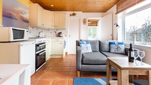 A compact kitchen with modern appliances, a grey sofa with nautical-themed pillows, and a dining area with a table and wine glasses. - Anchors Away - One Bedroom Cottage, Sleeps 2 (Downderry)