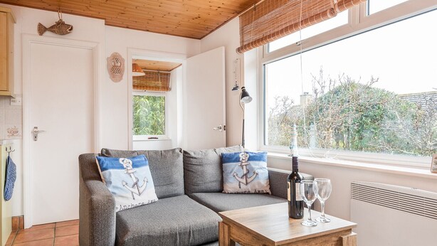 A cozy room with a grey sofa, two anchor pillows, a wooden table with a bottle and glasses, a window with a view of trees, and a ceiling fan. - Anchors Away - One Bedroom Cottage, Sleeps 2 (Downderry)