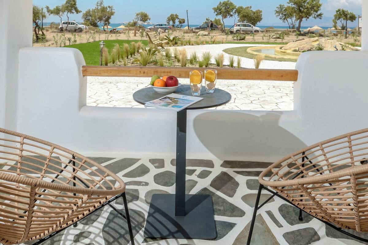 A terrace with a table, two wicker chairs, and a view of the beach and vehicles.