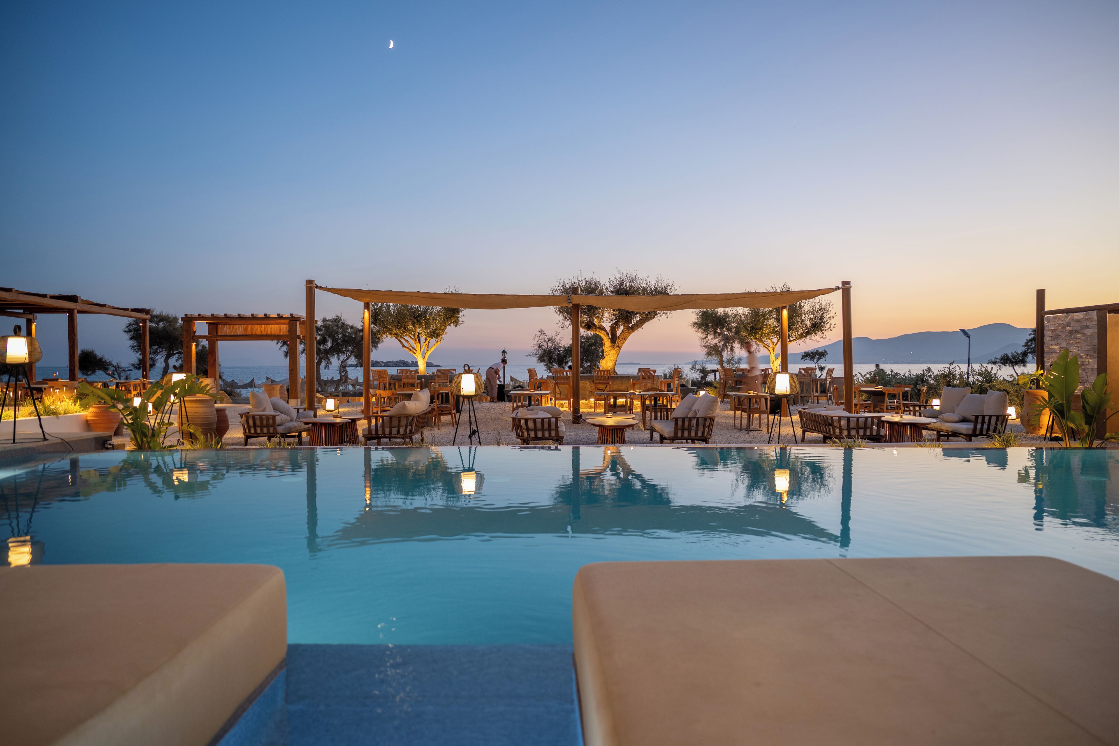 Un área junto a la piscina con sillones reclinables y mesas, una pérgola y vistas al mar y las montañas.