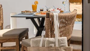 A dining table with breakfast items and a glass of orange juice, a wicker chair, and a bag with a label reading "Glaronissi Beach Luxury Suites & Apartments". - Glaronissi Beach (Naxos)