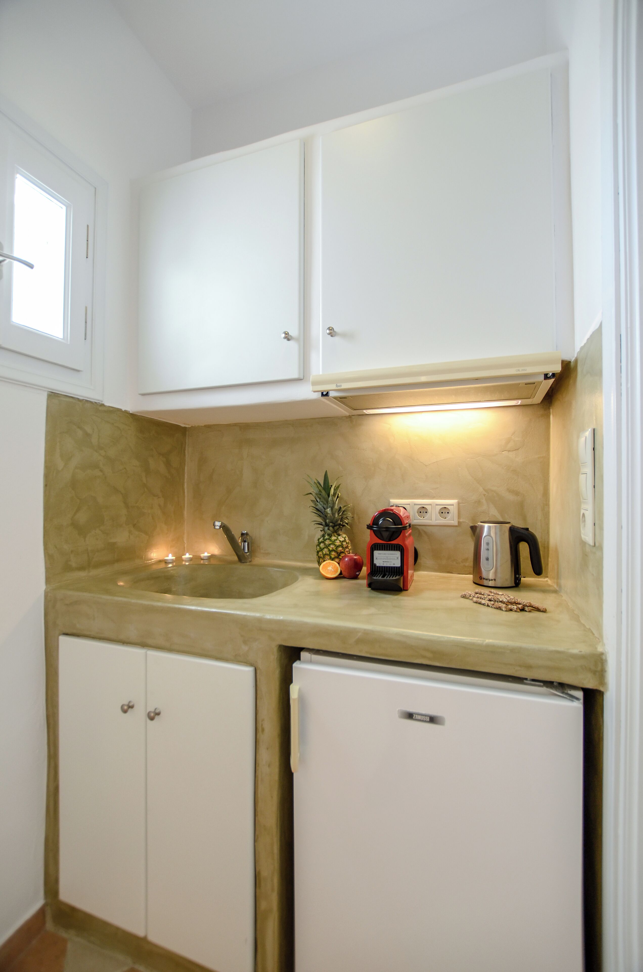 A compact kitchen with white cabinets, a countertop, a sink, a small refrigerator, a coffee maker, and a fruit bowl.