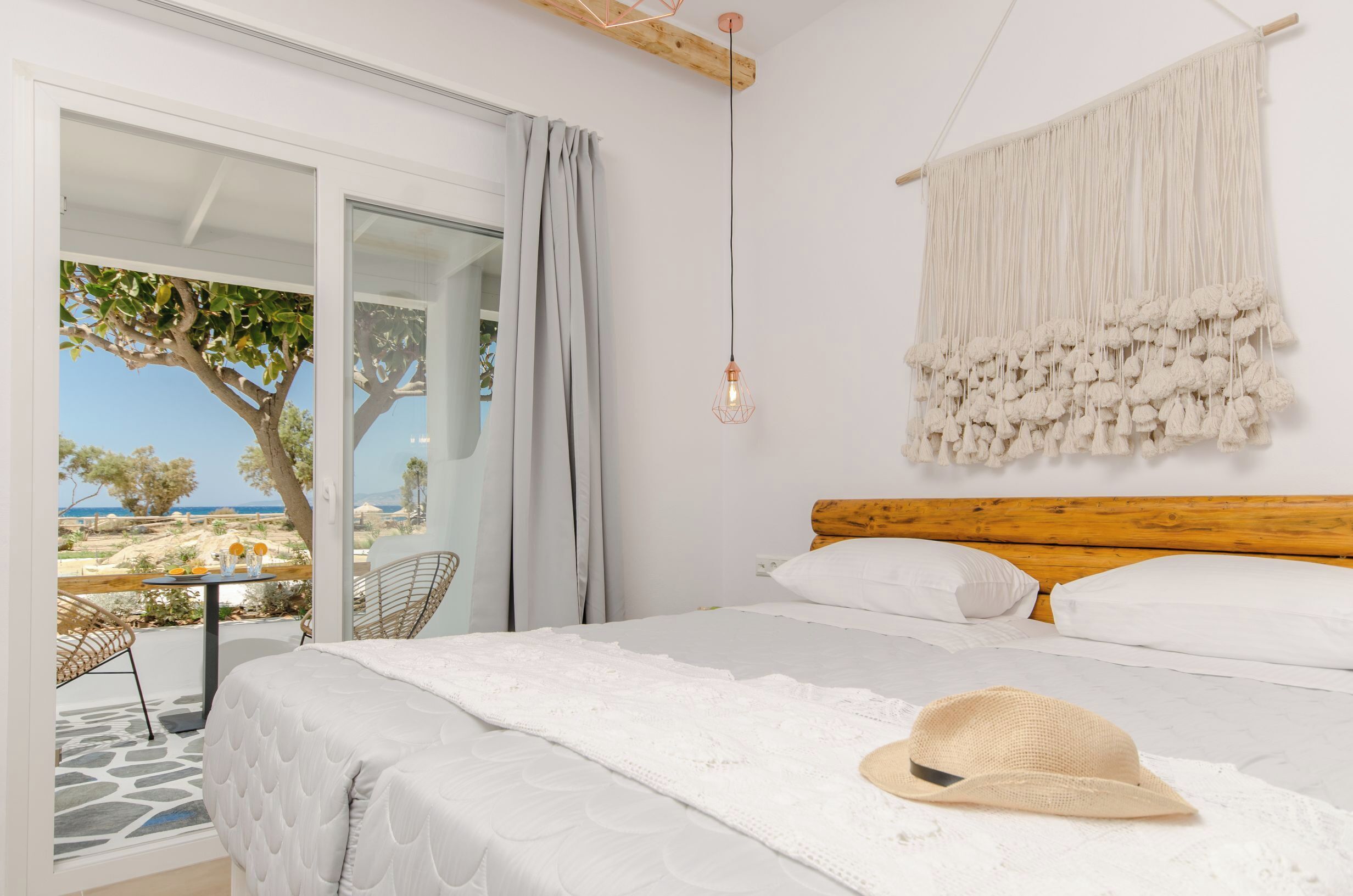 Une chambre avec un lit, une décoration murale en macramé suspendue et une vue sur la plage, visible à travers une porte coulissante.
