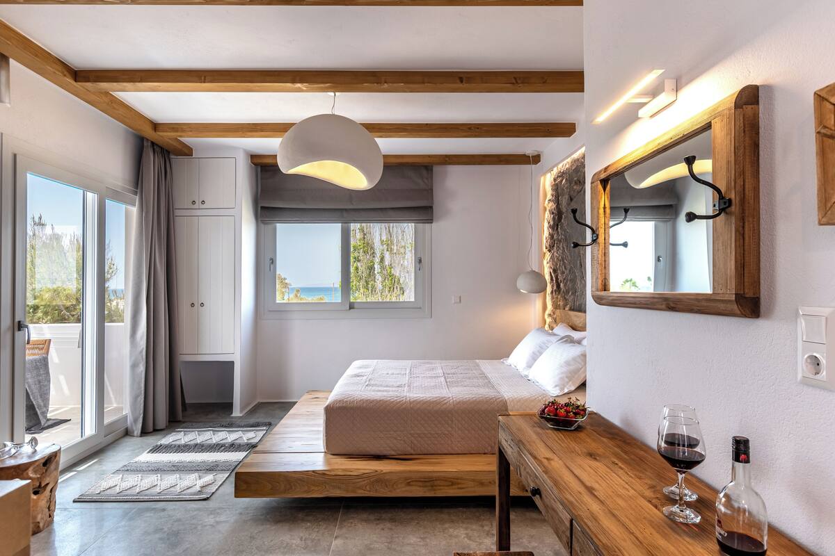 A bedroom with a bed, wooden beams, a hanging light, a window with a view, a mirror, and a bottle of wine on a table.