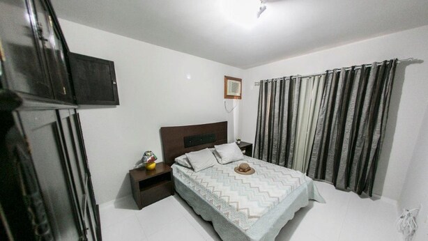 A bedroom with a bed, a nightstand, a TV mounted on the wall, and a window with curtains. - Casa Terceira avenida - 10 pessoas (Itapema)