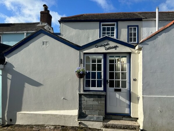 A cottage named 'Crumpet Cottage' with a white exterior, a blue door, and a small stone step leading to the entrance.