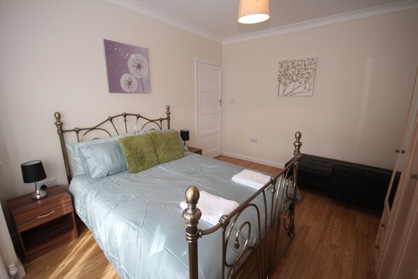A bedroom with a metal-framed bed, a nightstand with a lamp, and a door leading to another room. - Deane House (Ruislip)