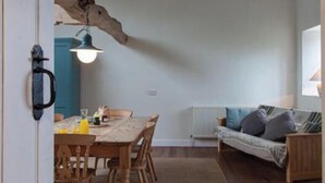 A dining area with a wooden table and chairs, a sofa, and a hanging light fixture. - Carneggan Barn - Three Bedroom Cottage, Sleeps 6 (Lanteglos)